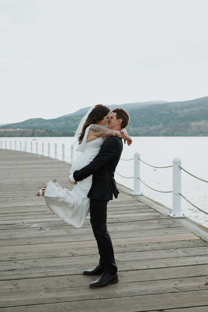 Groom lifting his bride for a kiss on a lakeside dock during their Okanagan wedding at The Laurel Packinghouse in Kelowna, BC, planned by a professional wedding planner.